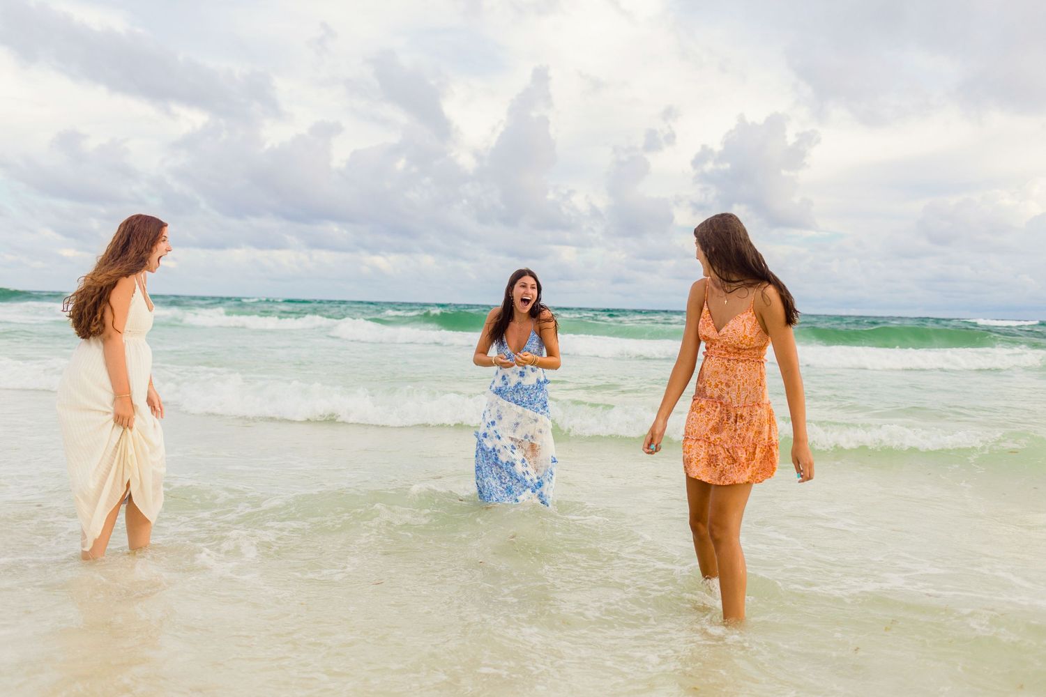 Three young women in colorful summer dresses laughing and wading in ocean waves on a tropical beach.