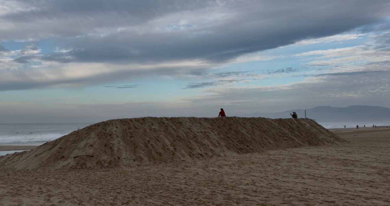Large sandy mound on a cloudy beach with figures standing on top, ocean visible in the background.