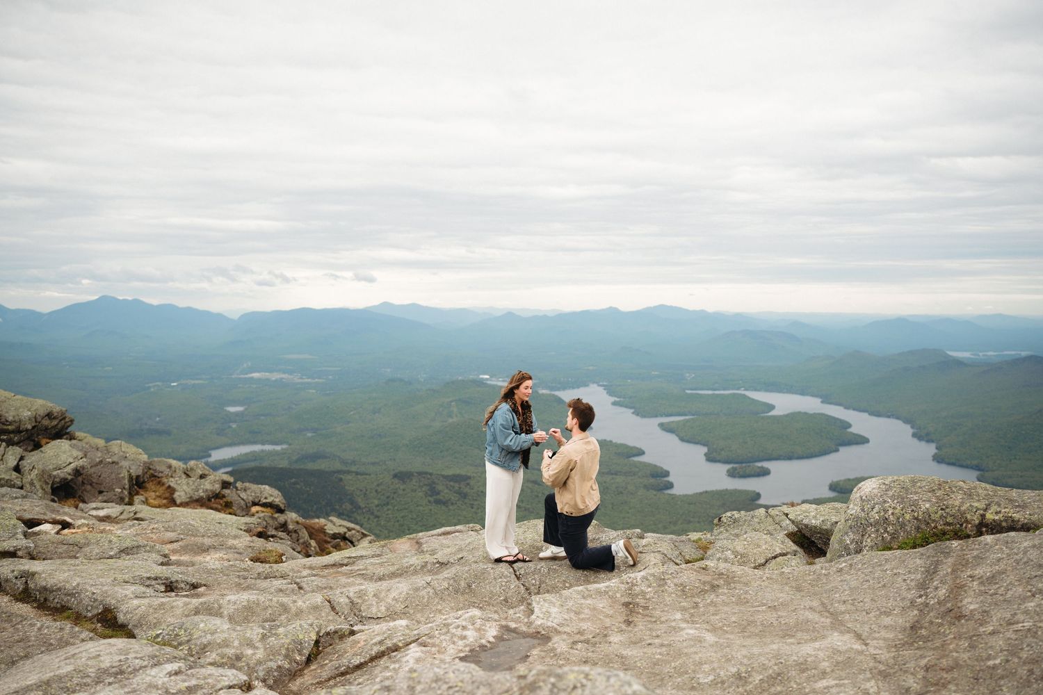 Man proposes on one knee atop a rocky mountain summit with sweeping views of green valleys and a winding river below.
