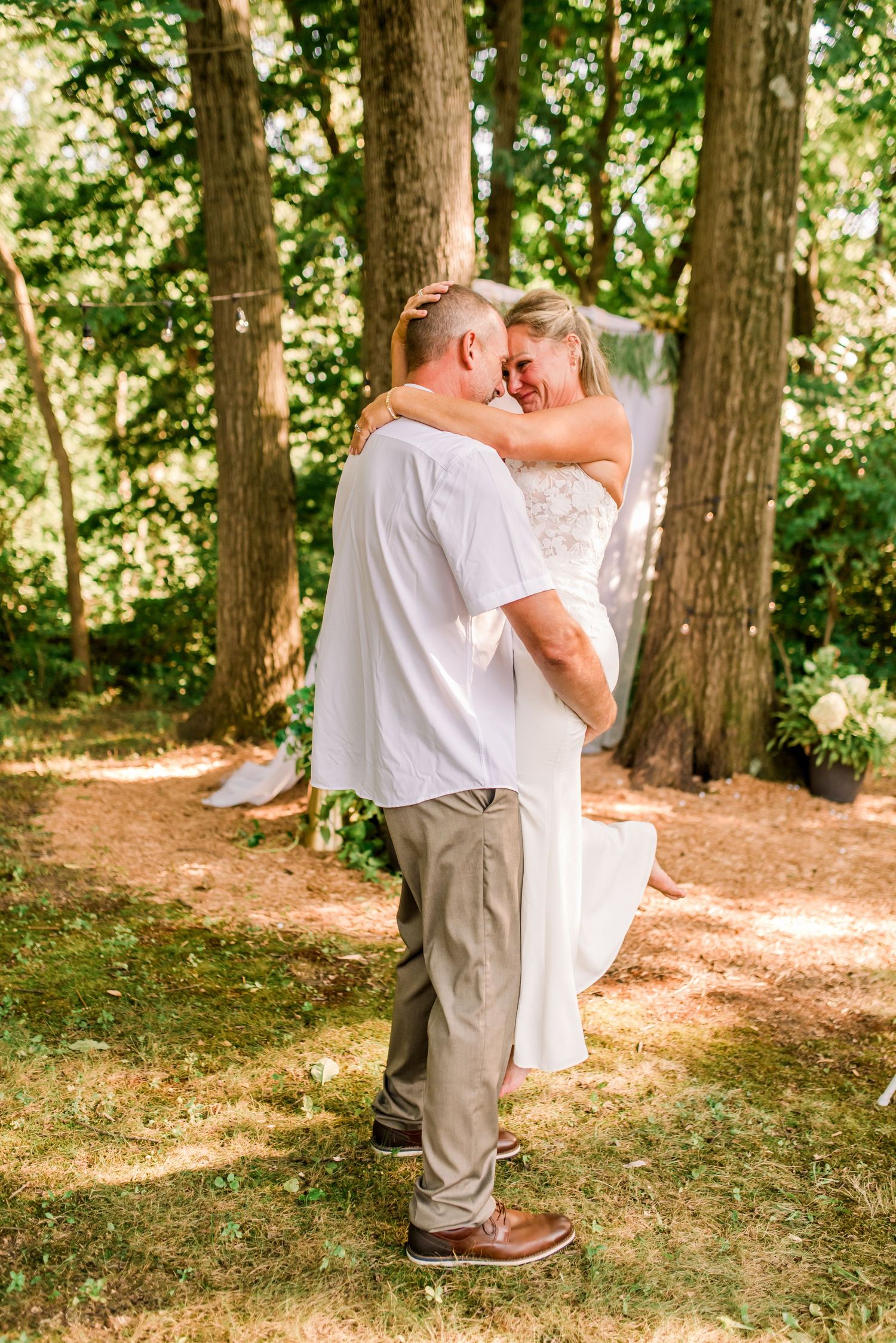 A joyful newlywed couple holding hands and walking outdoors among trees, bride in white gown, groom in white shirt and khakis.