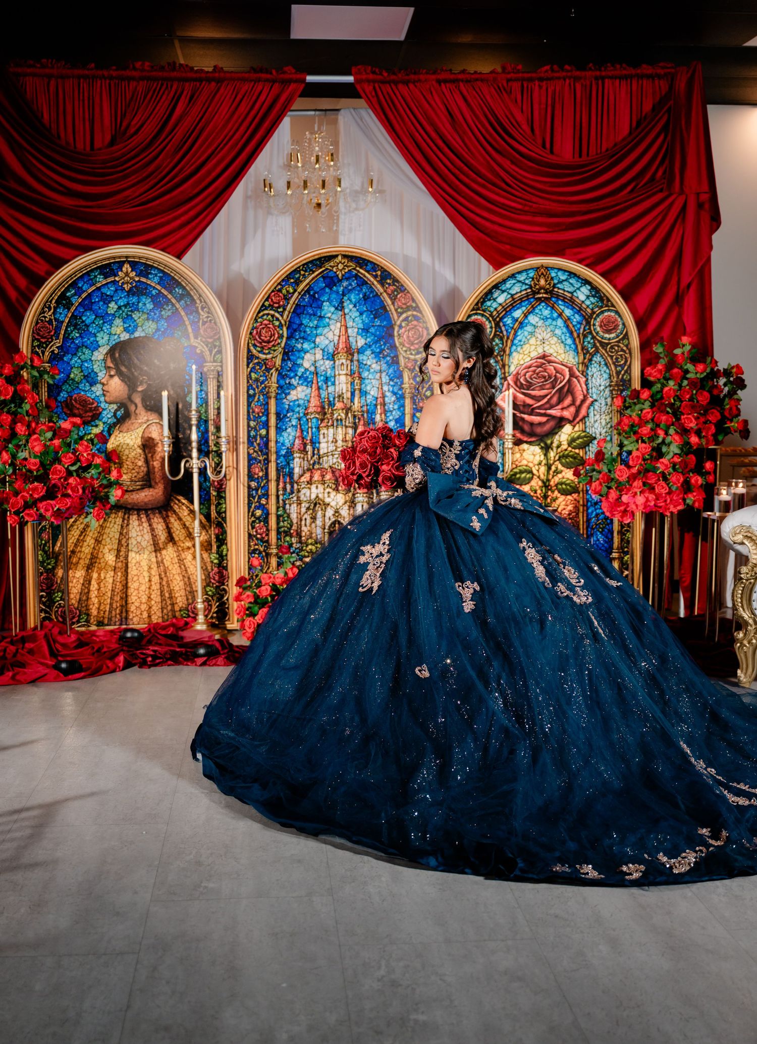 Young woman in a navy blue embroidered quinceañera ball gown holding red roses in front of fairytale castle stained glass panels.
