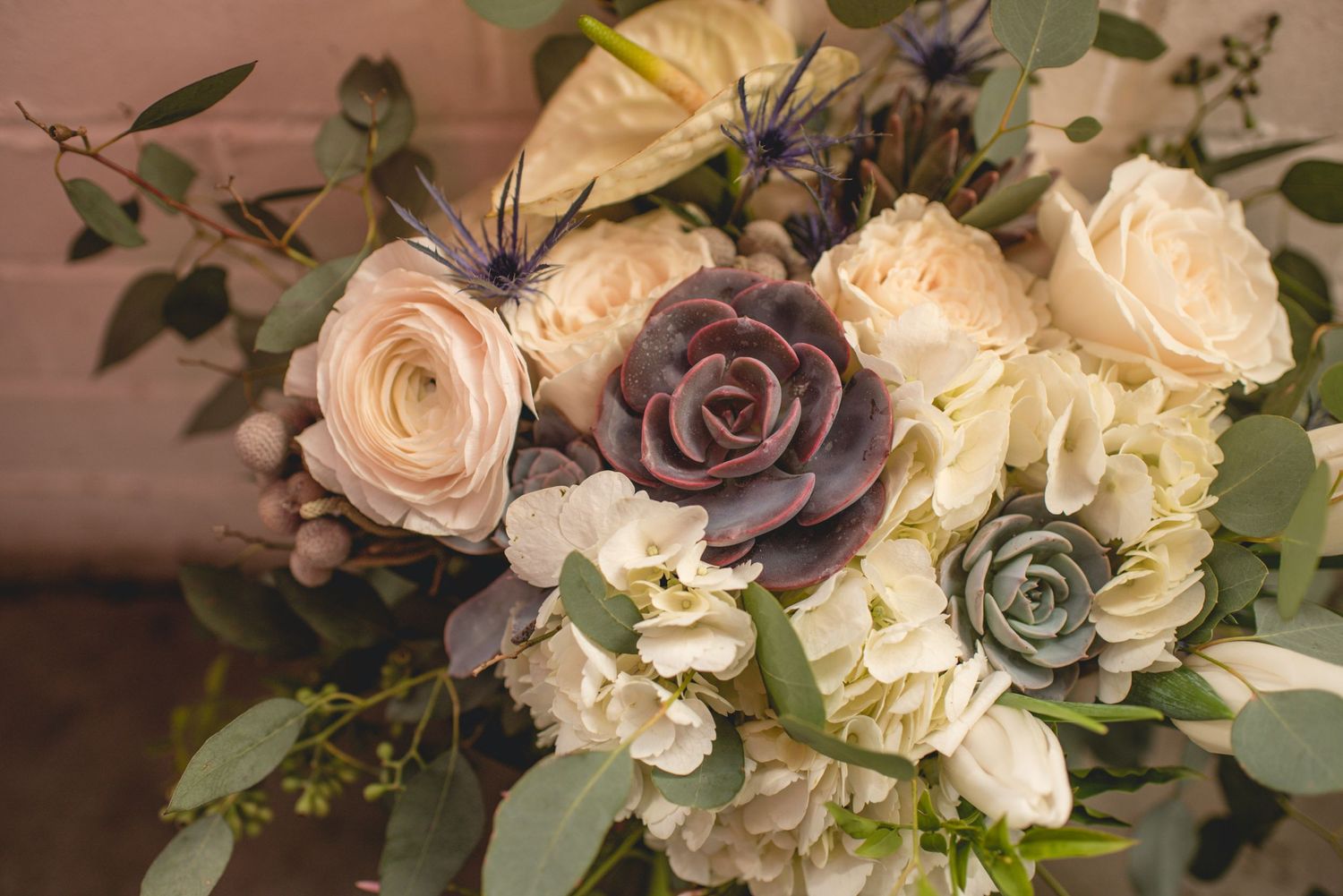 Elegant floral bouquet featuring cream roses, purple blooms, and greenery against a soft blush background.