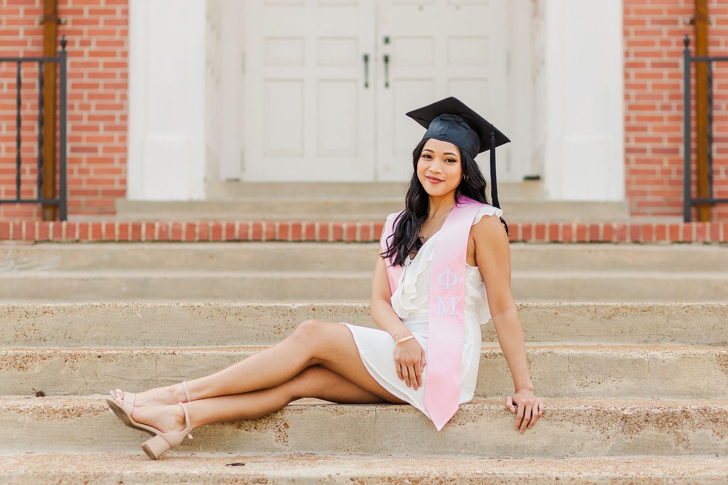 A graduate in white dress and cap poses elegantly on university steps.