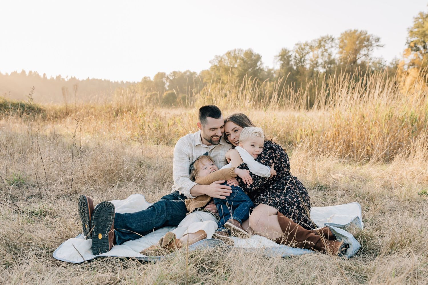 Family of four sitting together on a blanket in a golden field, sharing a warm embrace during an outdoor photo session.