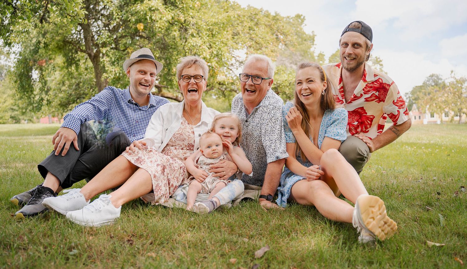 Multiple generations of family members relax together on grass in a sunny park.