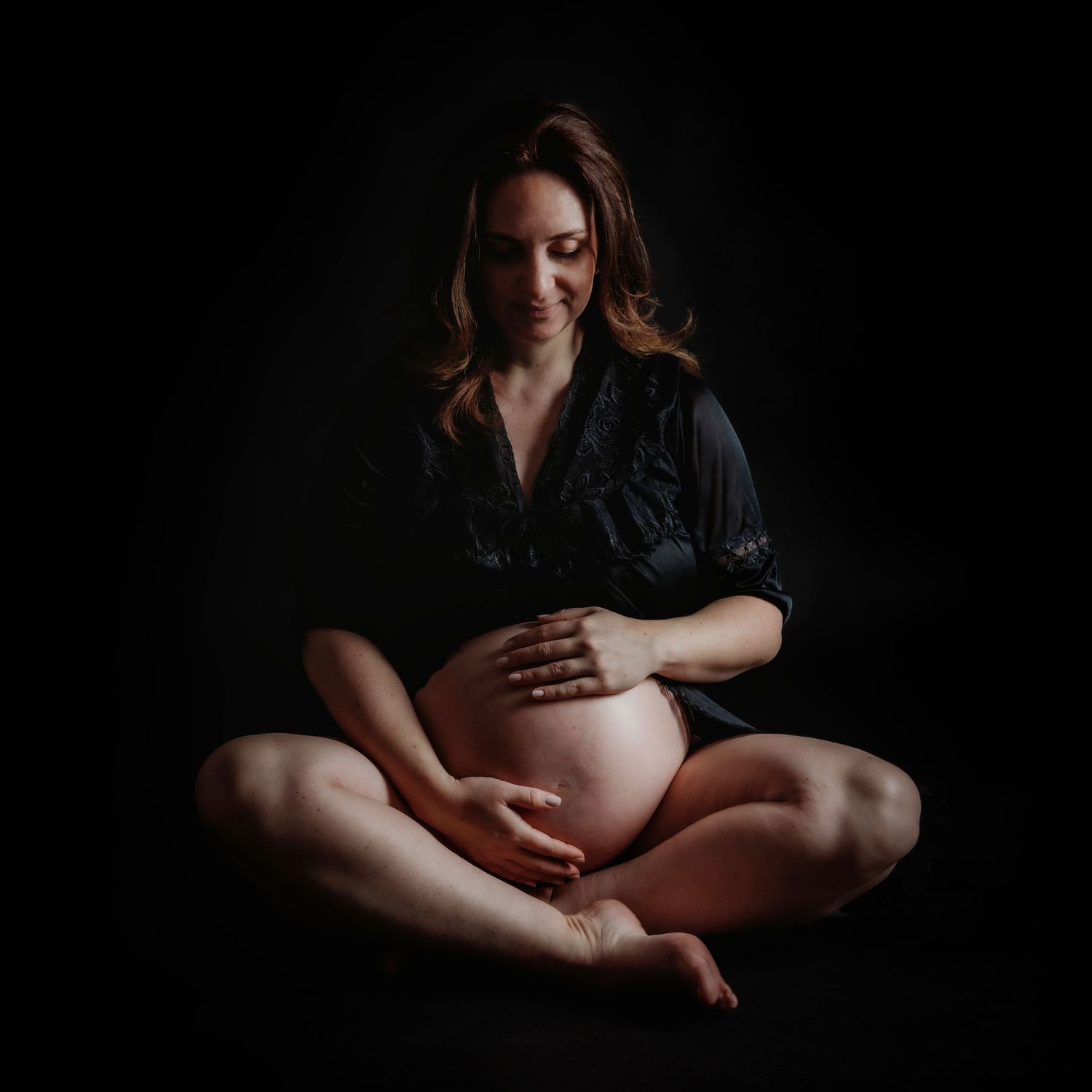 Artistic maternity photo of an expectant mother in black dress sitting cross-legged against dark background.