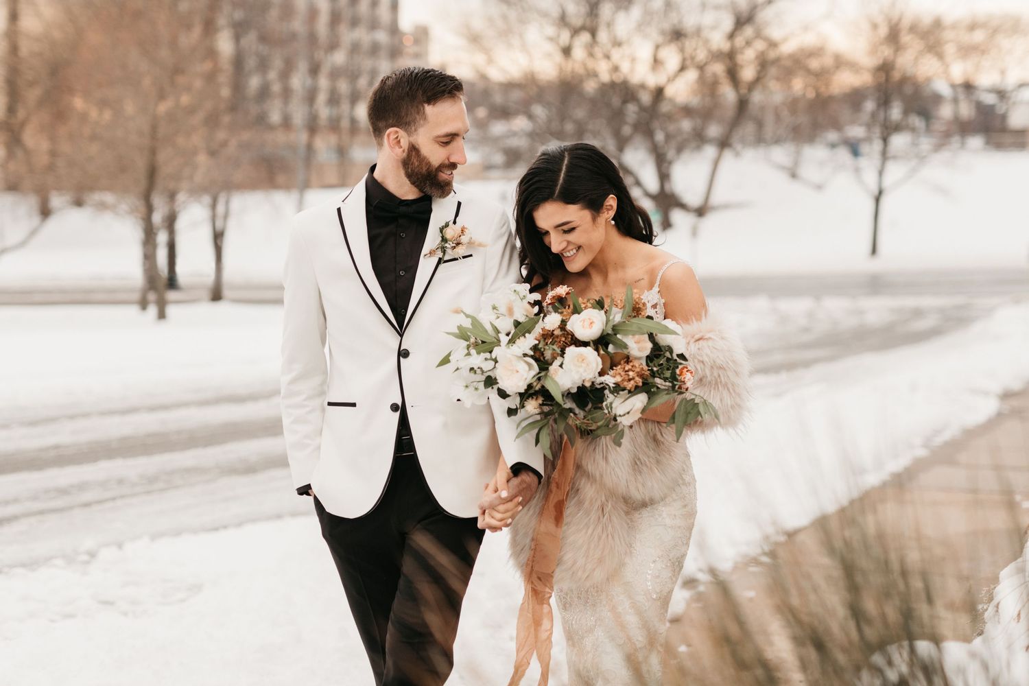 Couple in winter wedding attire walking along snowy path with bridal bouquet.
