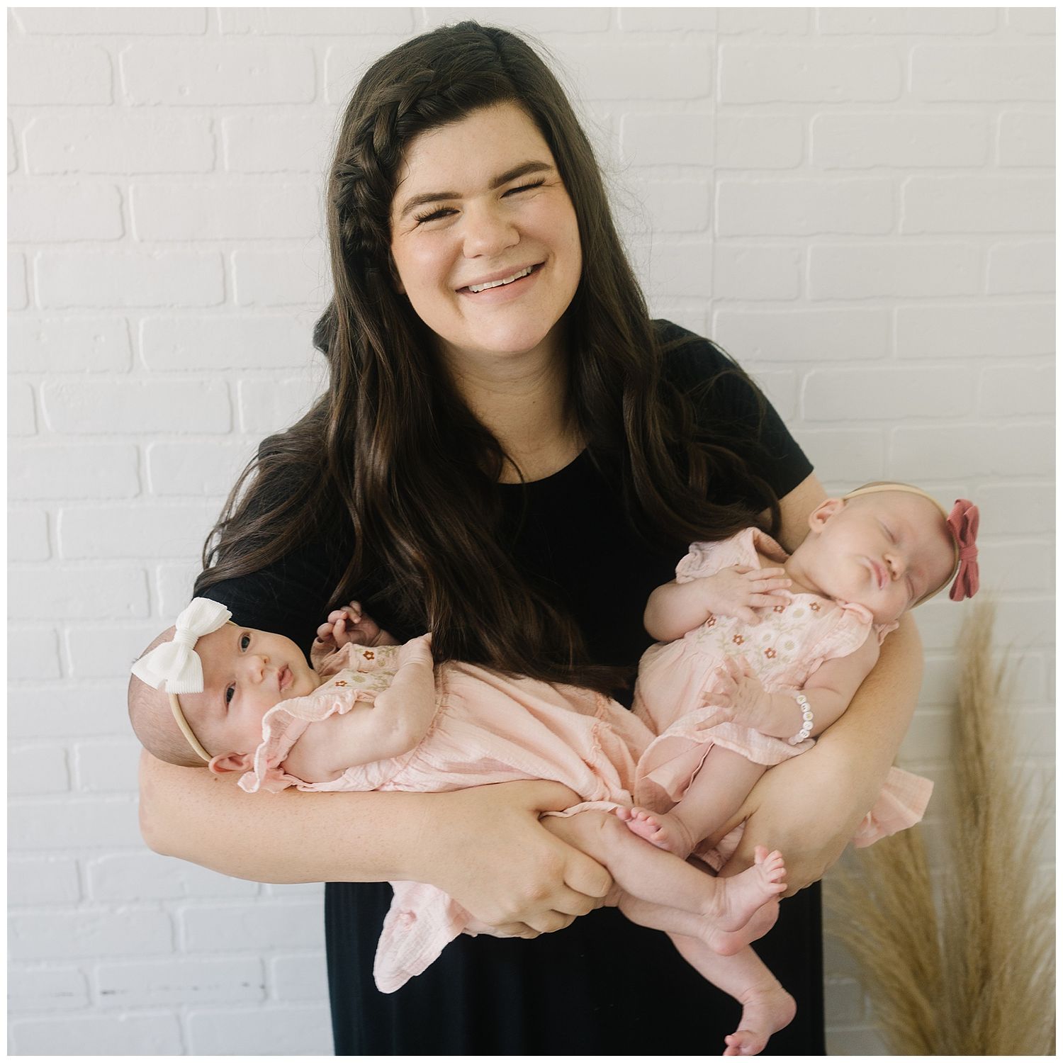 Woman in black holding twin babies in matching pink outfits against white brick wall.