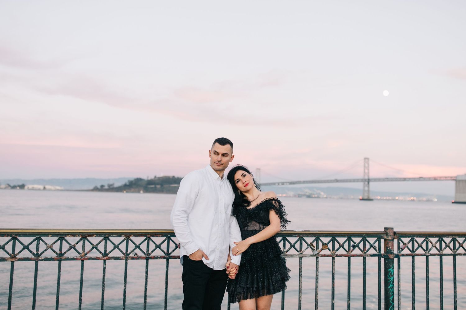 A couple poses on a pier at sunset with the San Francisco Bay Bridge visible in the background.