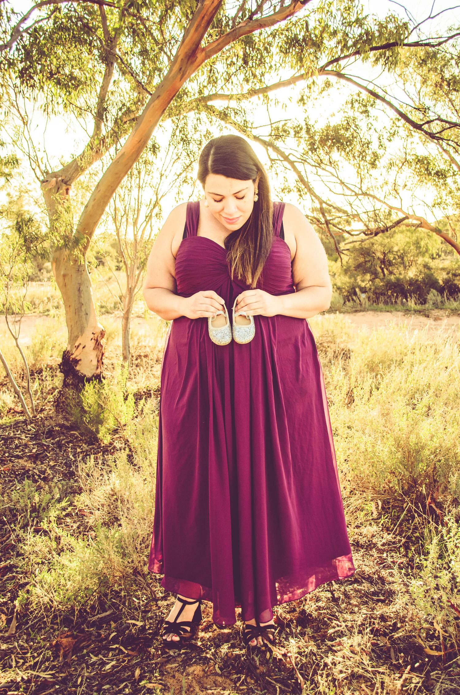 A person in a burgundy formal dress stands in a sunlit eucalyptus grove.