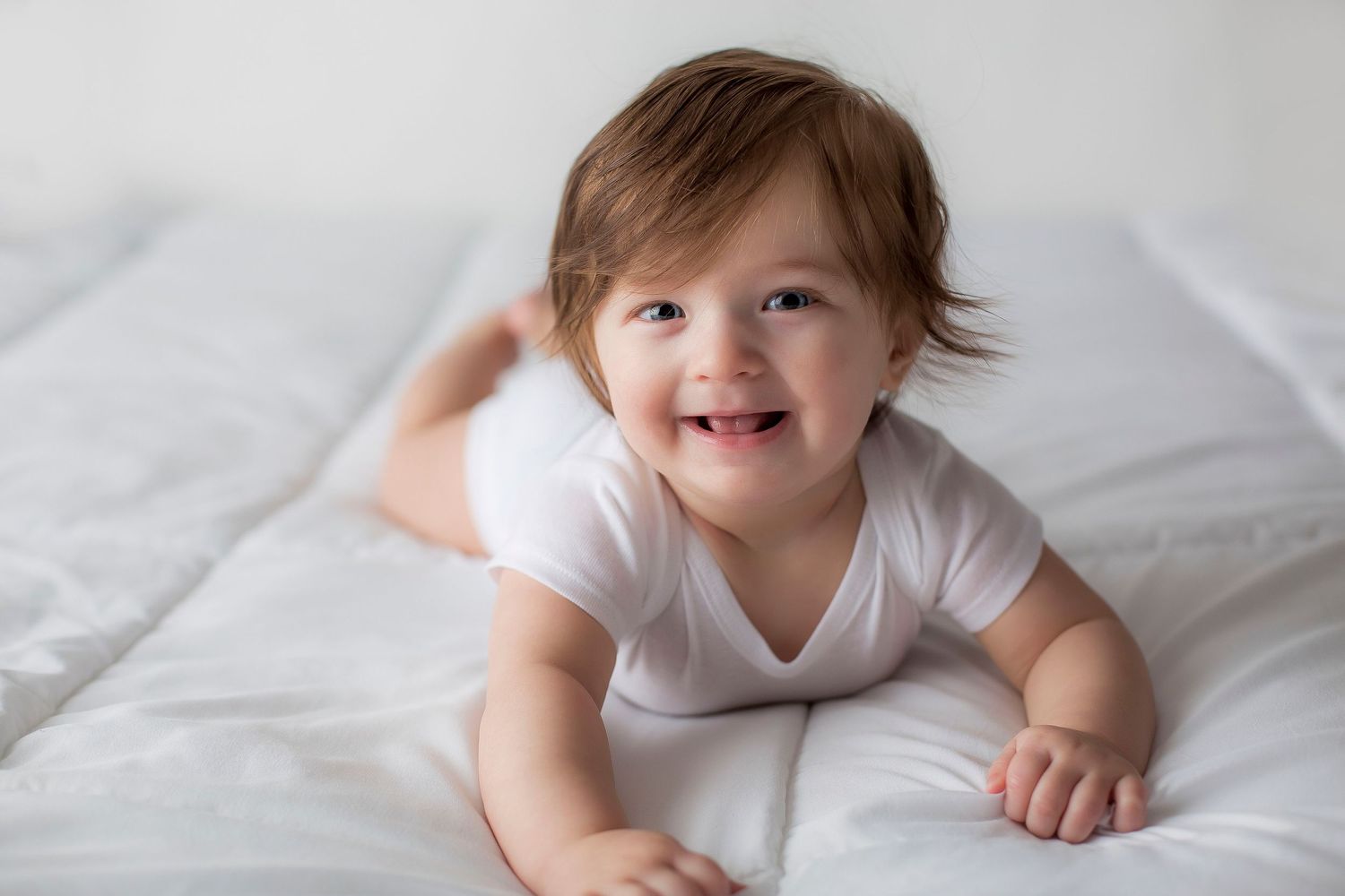 6 month old baby boy laying on a white blanket doing tummy time smiling at the camera.