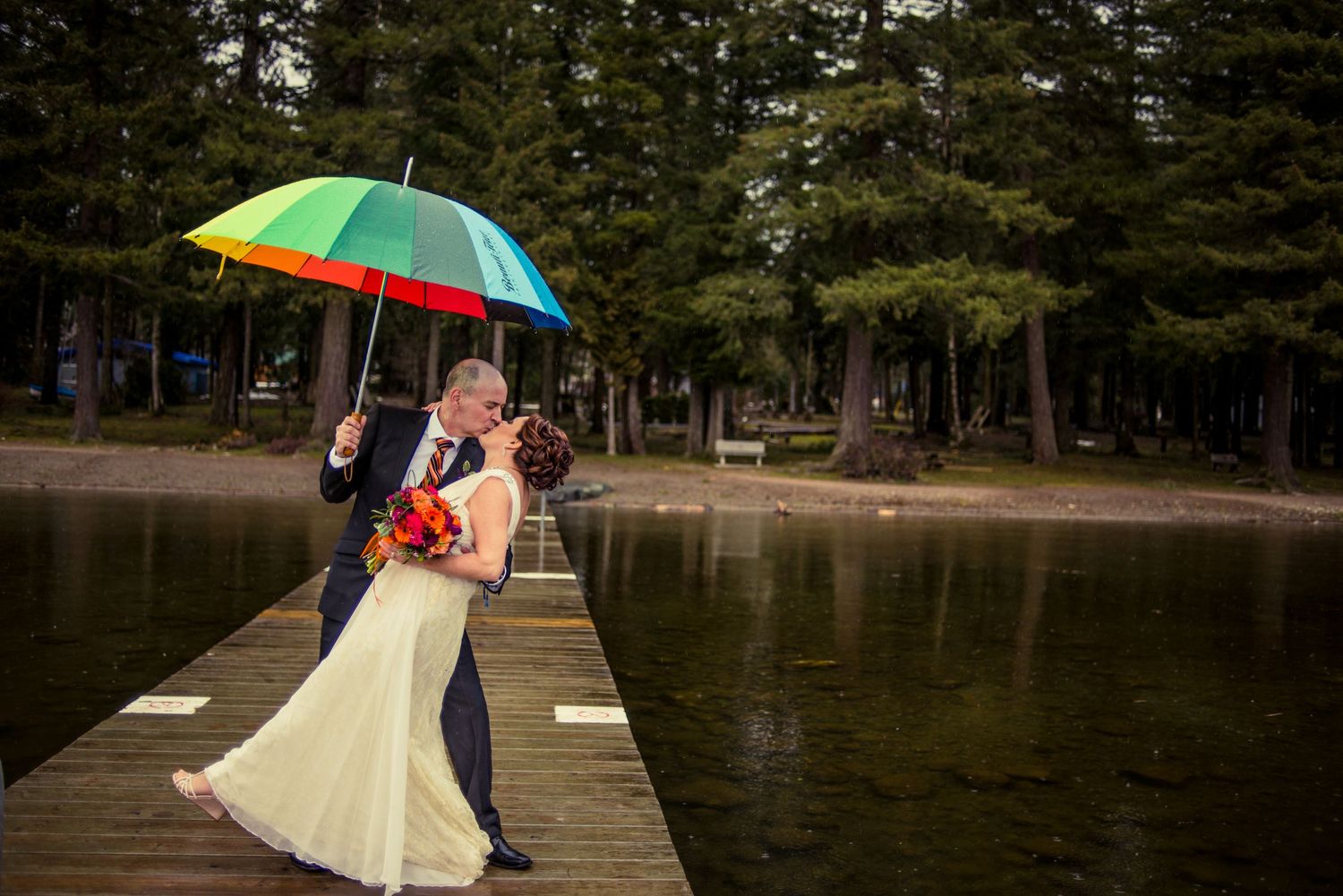 Couple embracing on a dock with colorful umbrella by a serene lake surrounded by pine trees.