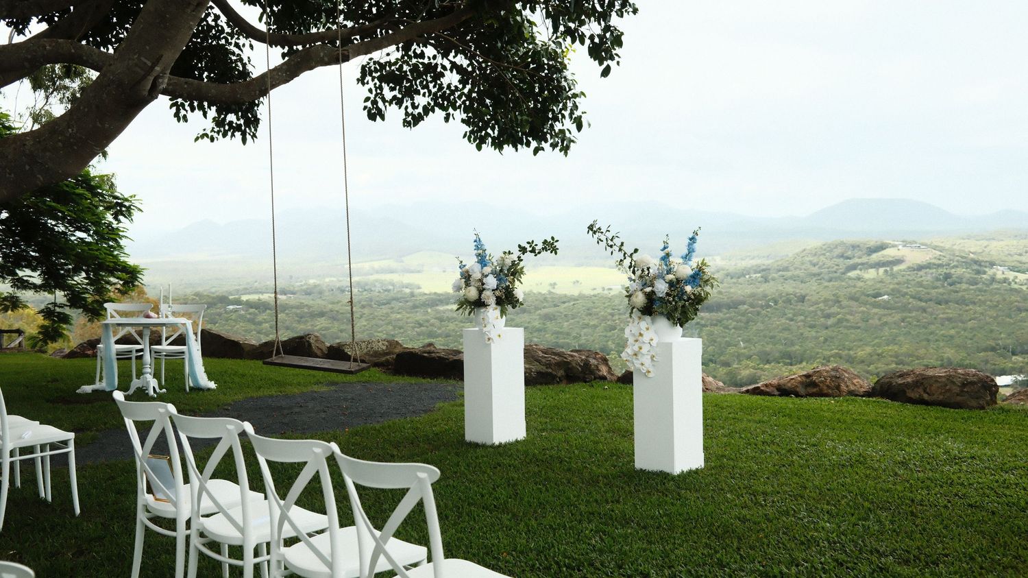Elegant outdoor wedding ceremony setup with white chairs, floral arch, and stunning valley views under a large tree.