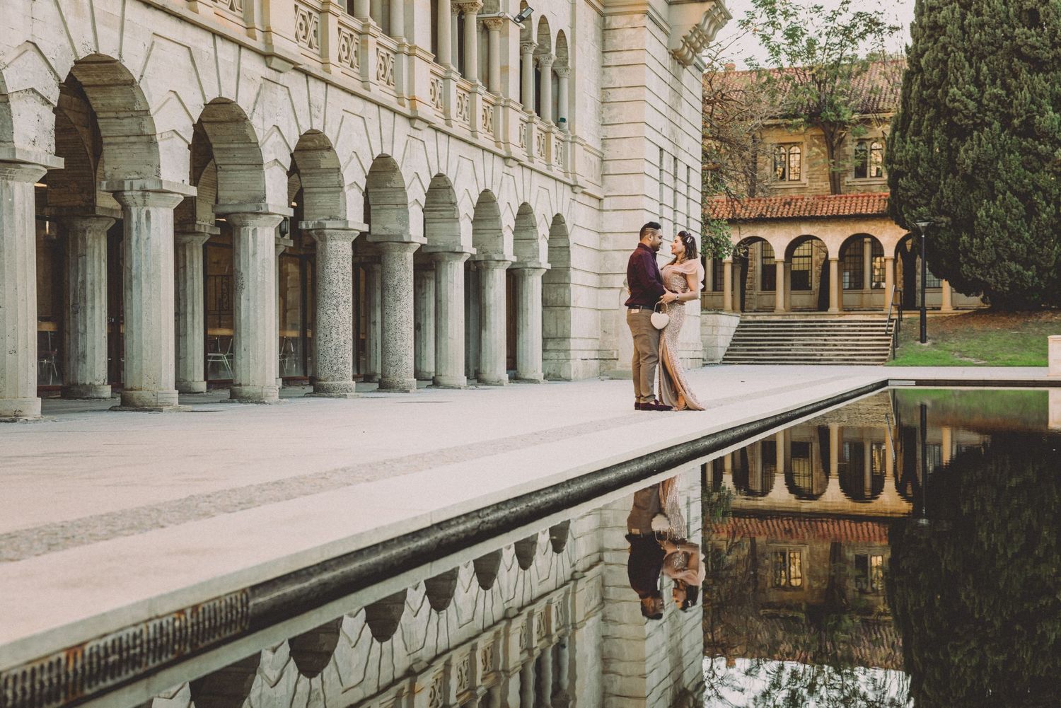 Couple embracing by a reflective pool at an elegant arched building, captured in a romantic engagement photo.