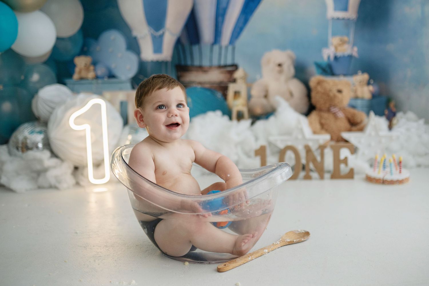 Baby sitting in bowl at first birthday cake smash with teddy bears, balloons, and number one decorations.