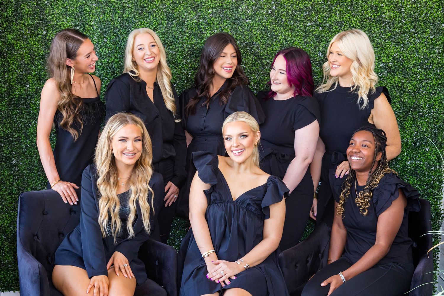 Eight women in matching black outfits pose together smiling in front of a green hedge wall backdrop.
