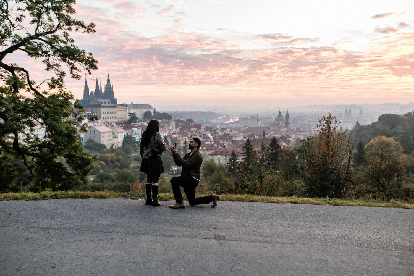 Man kneels on one knee proposing to a woman at a scenic hilltop overlook with a castle and city skyline in the background at sunset.