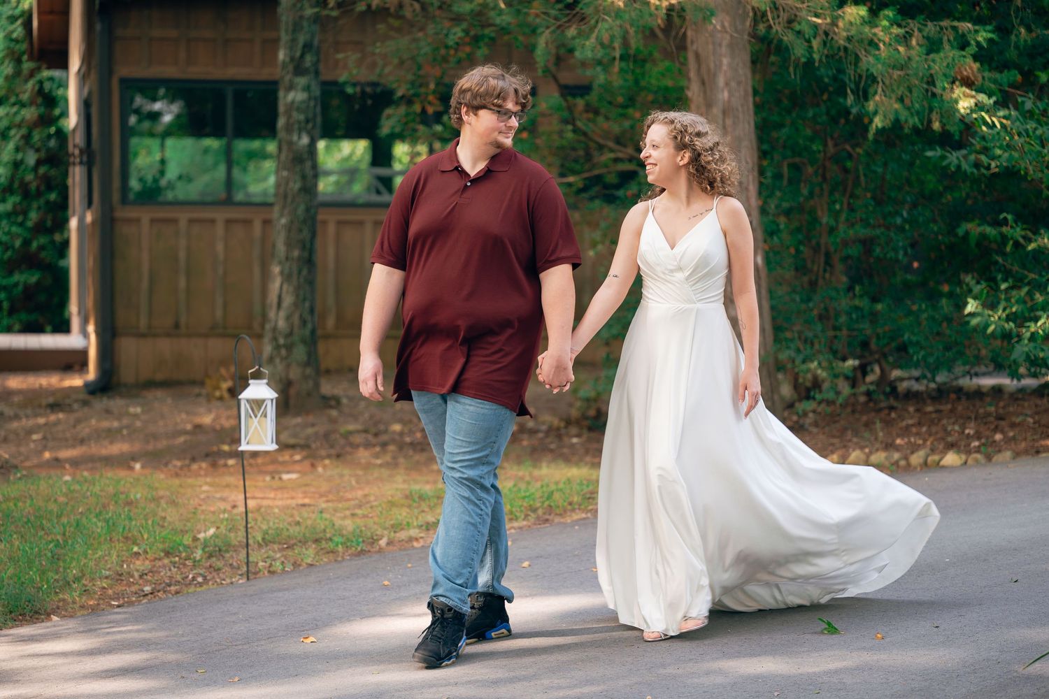 Bride in white gown and groom in maroon shirt walk hand in hand on a tree-lined path outdoors.