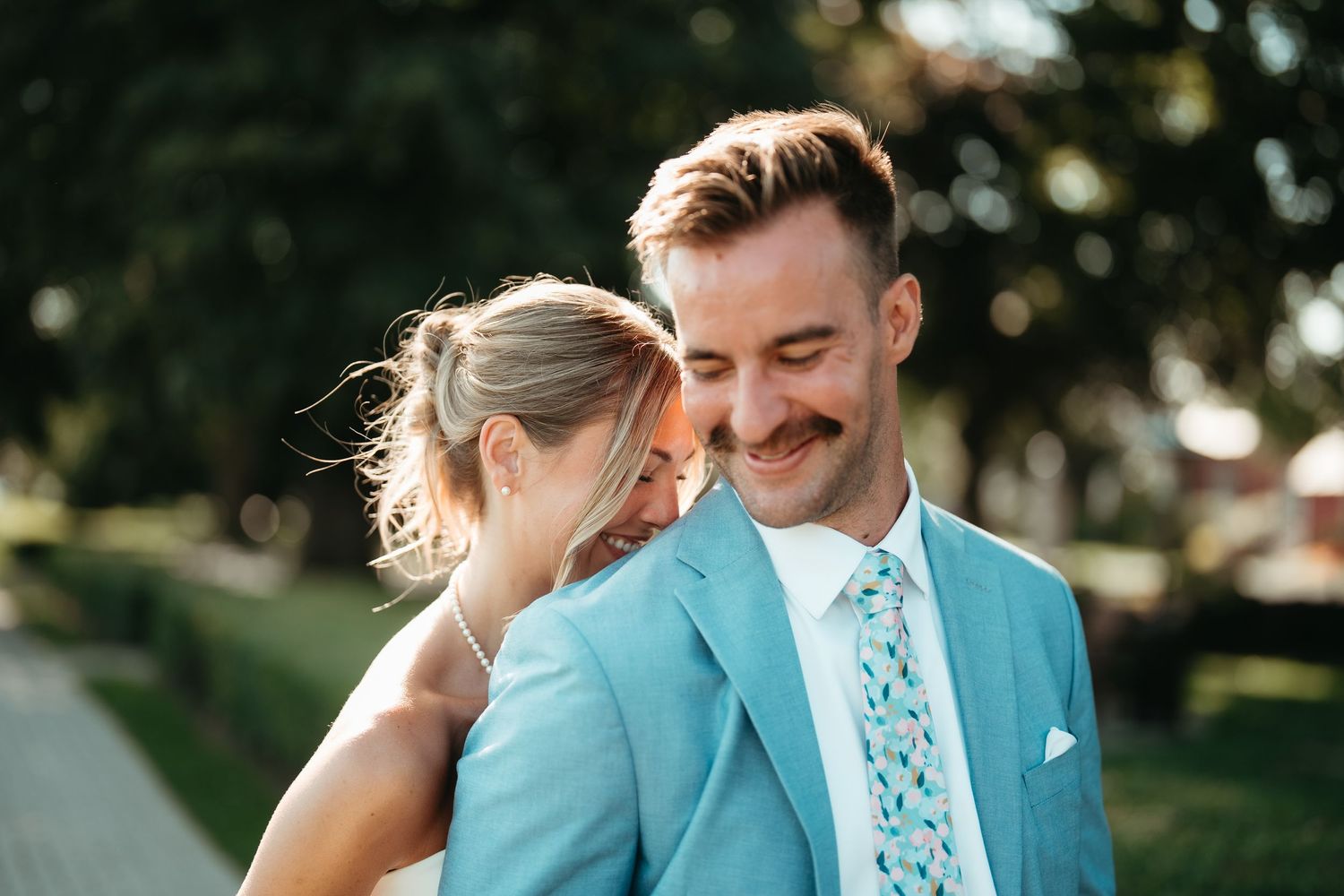 Couple in love smiling outdoors, groom in light blue suit with floral tie, bride in white dress leaning on his shoulder.