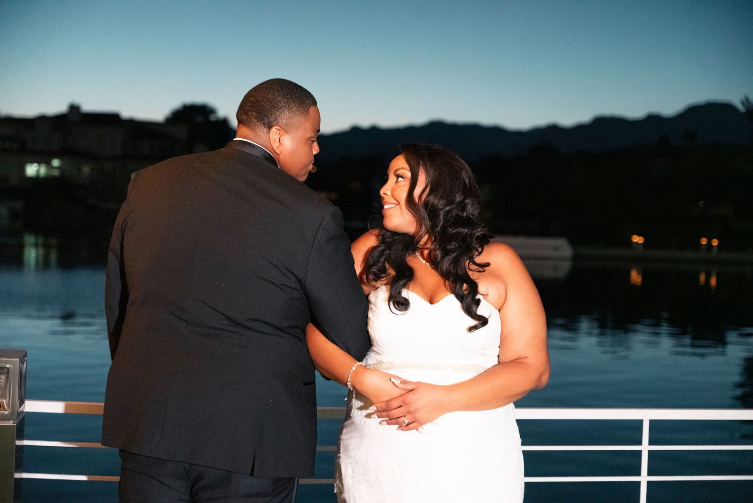 Couple shares romantic moment on waterfront deck at dusk, bride in white gown and groom in black suit with mountains behind.