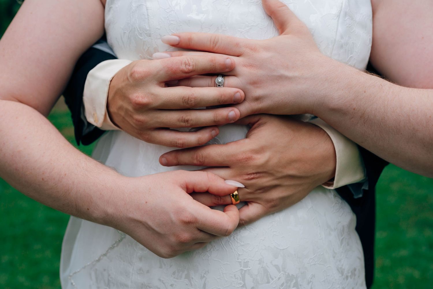 Close-up of a newlywed couple embracing, showing wedding rings on their hands against a white bridal gown.