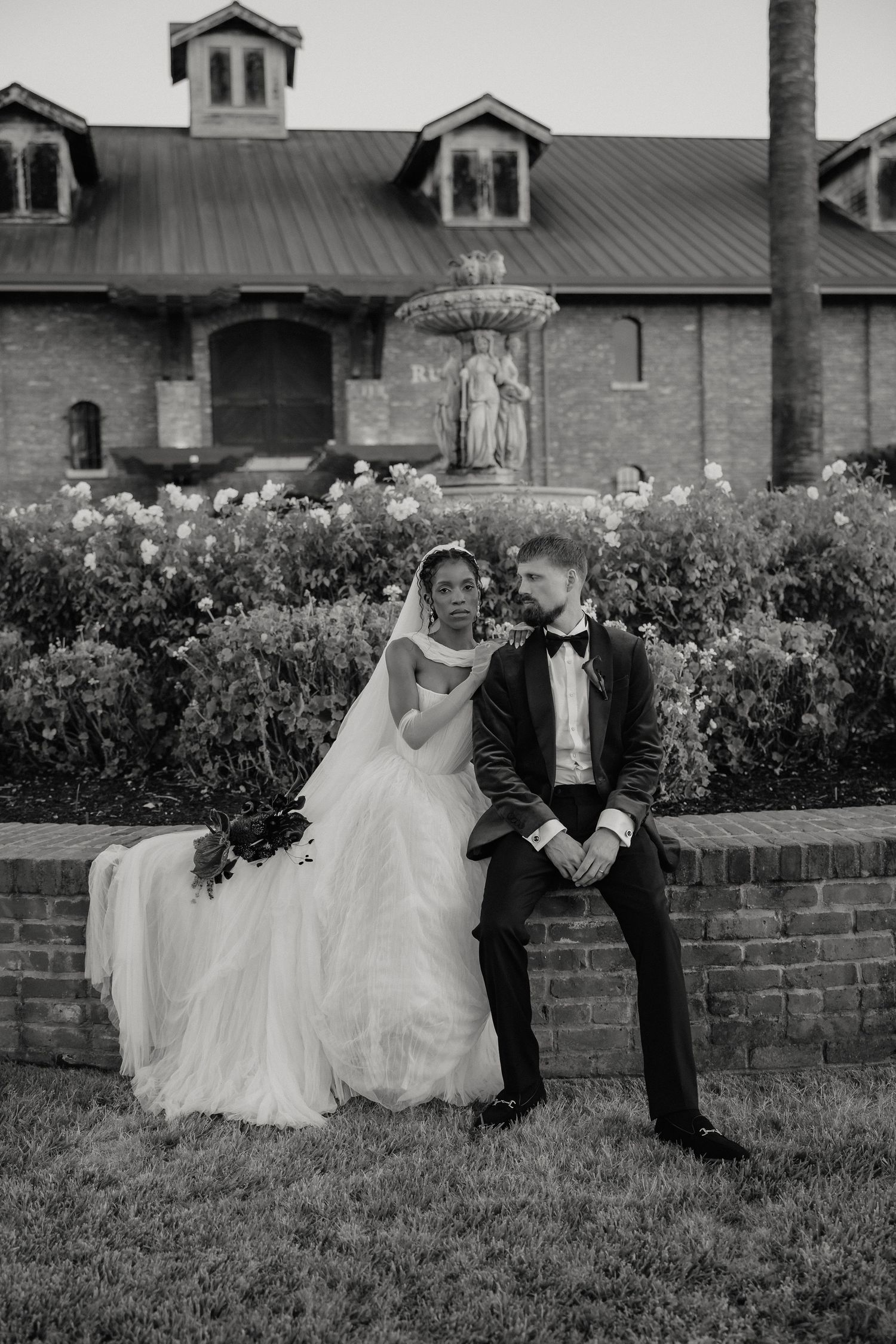 Black and white photo of bride and groom sharing an intimate moment under flowing wedding veil in outdoor field.