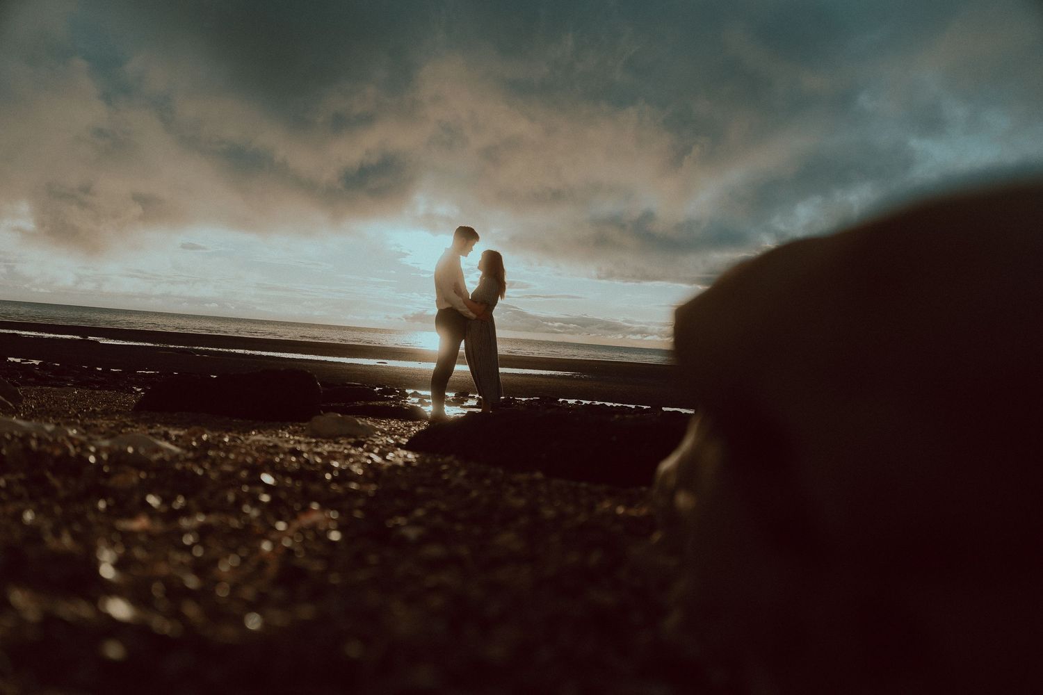 Couple embracing at sunset on a dramatic beach with moody cloudy sky and golden light reflecting on wet sand.
