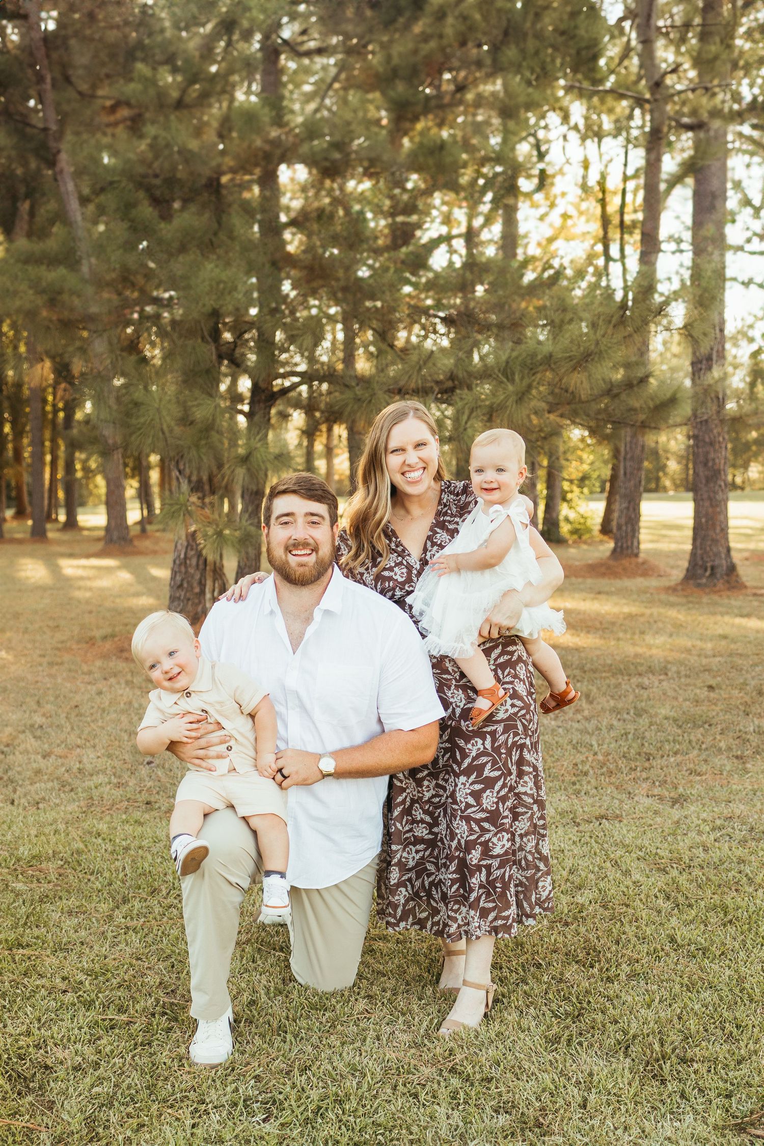 Happy family of four with two babies poses outdoors in pine tree park during golden hour sunset.