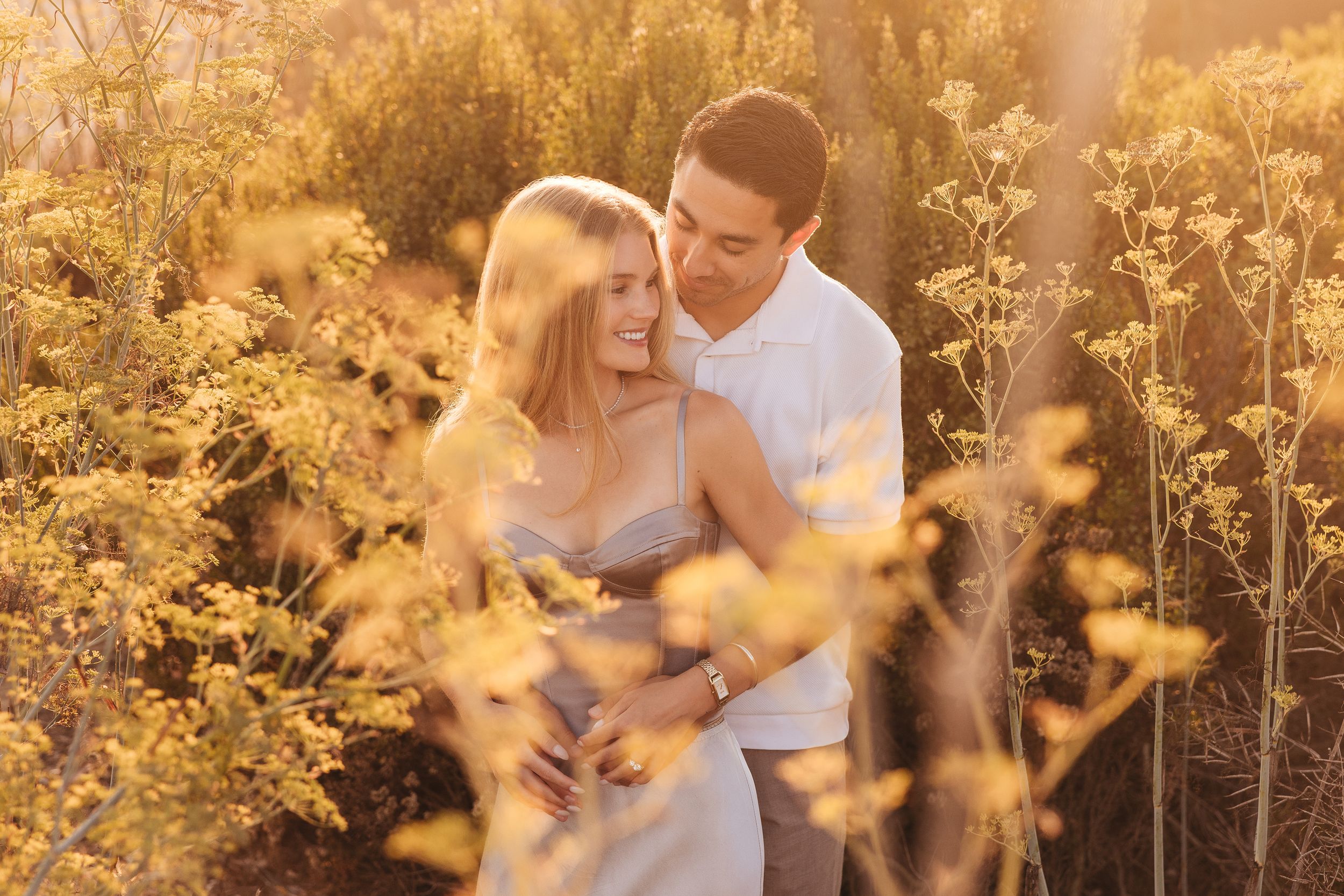 Couple embracing romantically in golden wildflower field during sunset, woman in gray dress, man in white shirt.