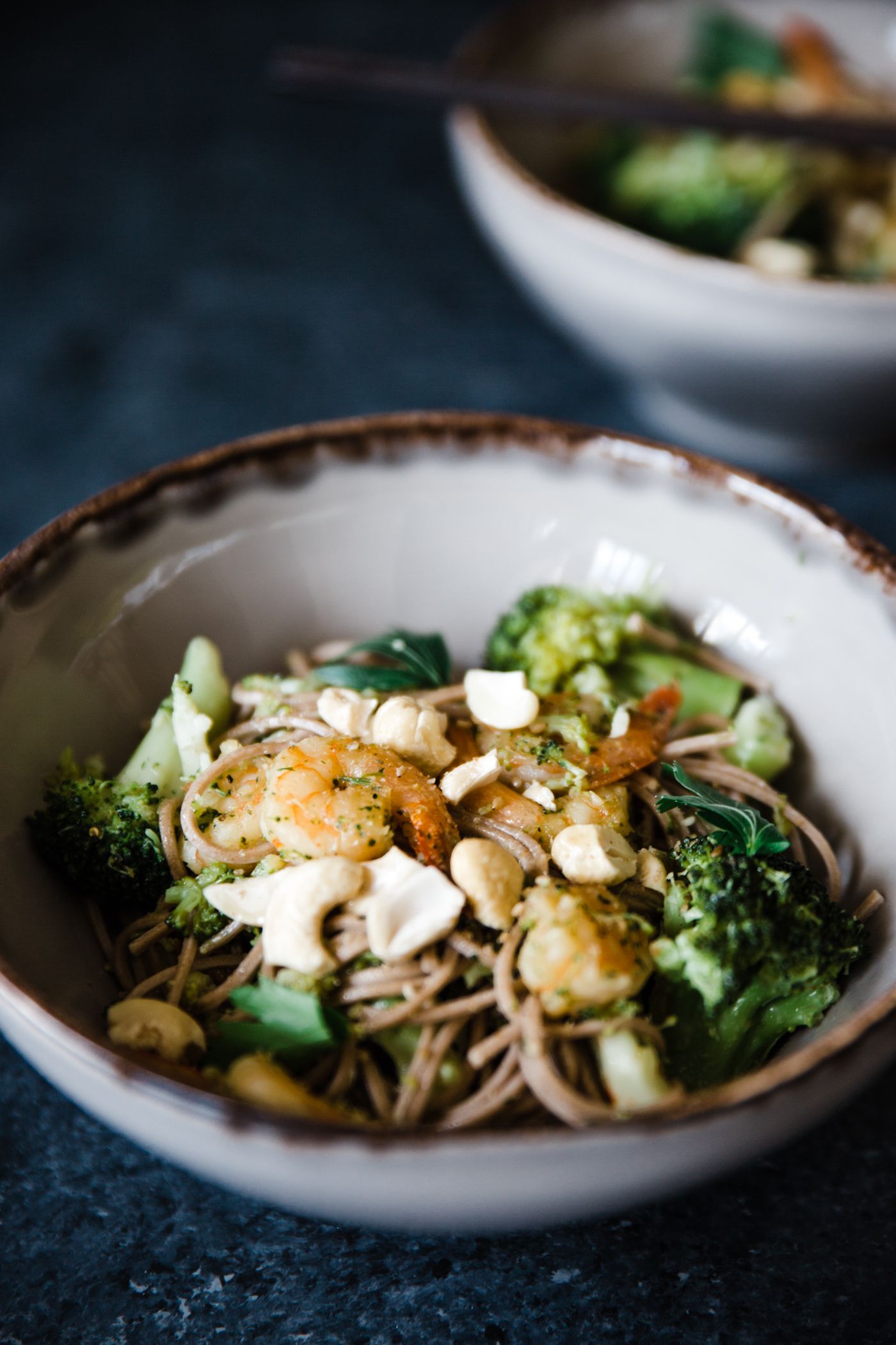 Bowl of soba noodles with broccoli, mushrooms and nuts in moody lighting.