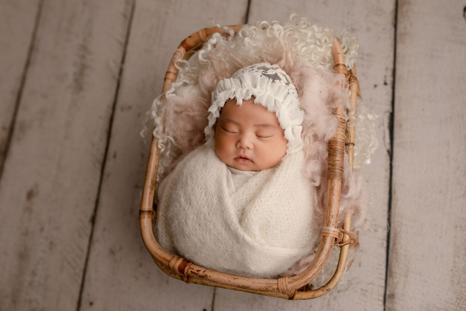 newborn baby girl wrapped in white knitted wrap on pink wool in bamboo basket on cream wooden planks