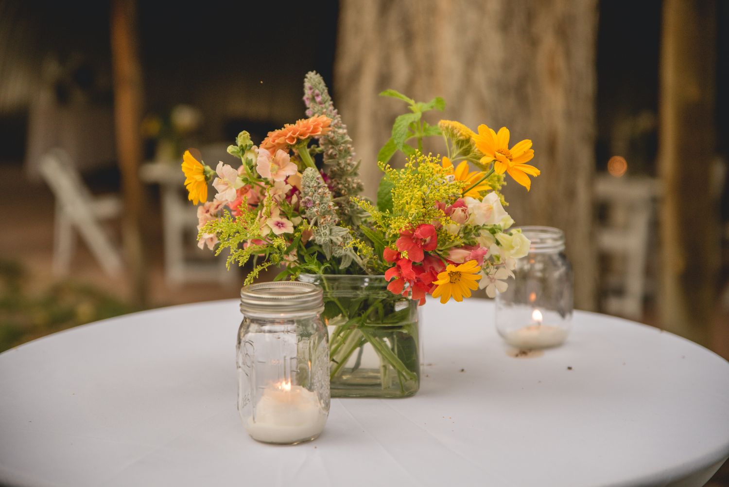 Colorful wildflower centerpiece in mason jar with candle on white table.
