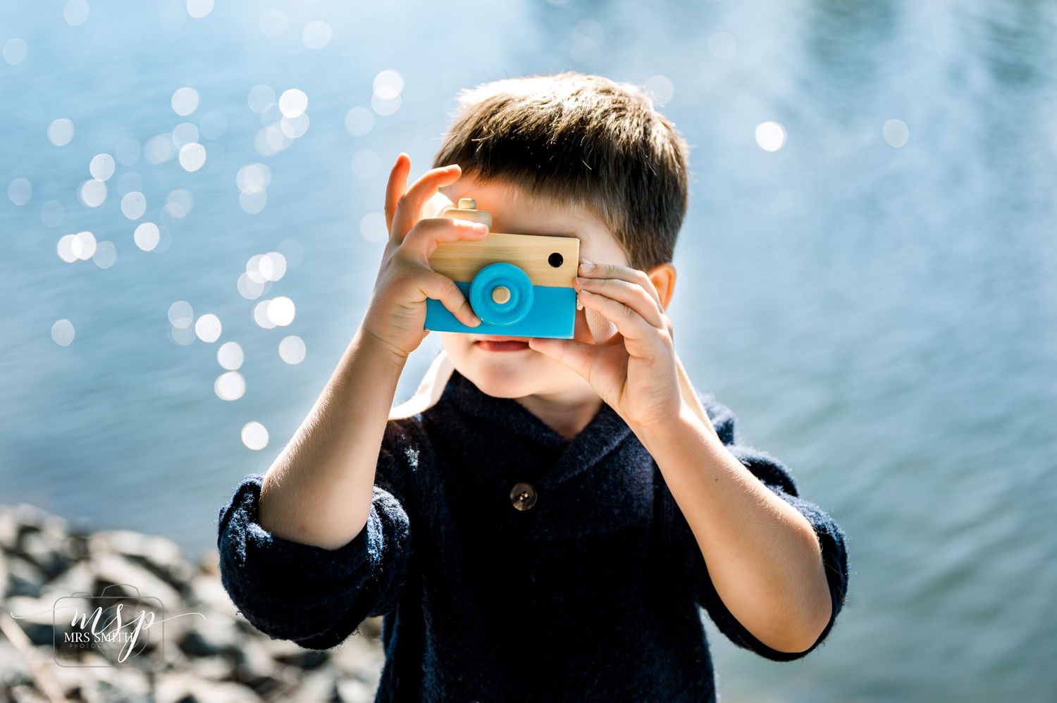 Young child holding a blue toy camera outdoors with bokeh lights in background.