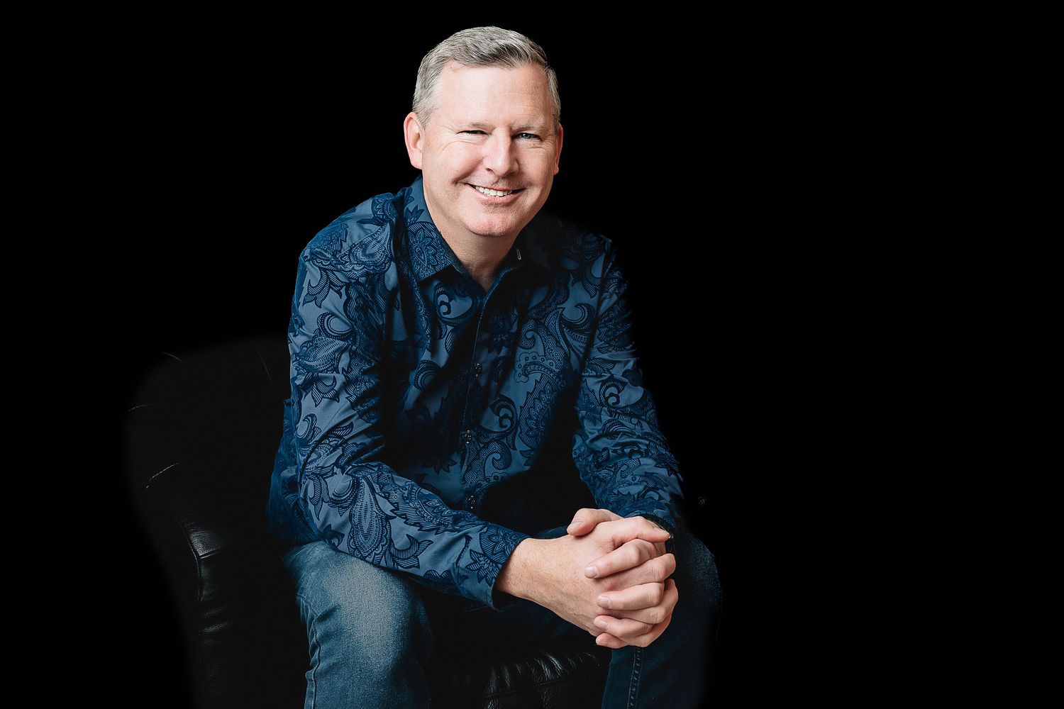 Professional headshot of a man wearing blue patterned shirt against black background.
