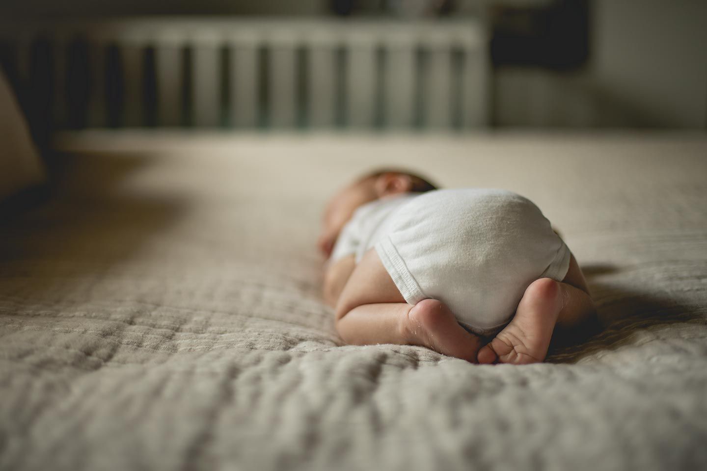 newborn baby laying on bed in a white onesie and feet tucked under body