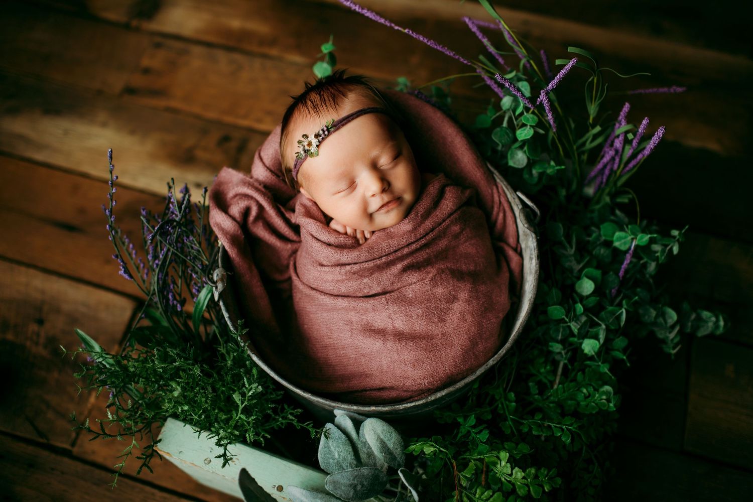 Sleeping newborn wrapped in brown swaddle surrounded by greenery in rustic setting.