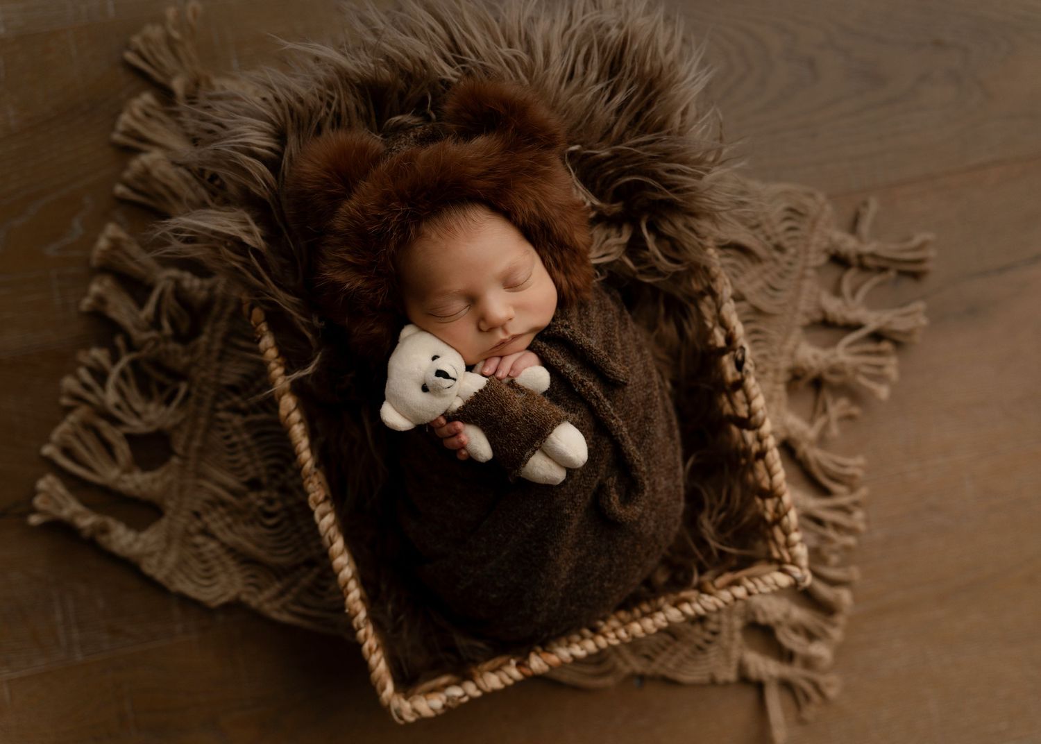Sleeping newborn in brown furry wrap holding stuffed toy in cozy basket.