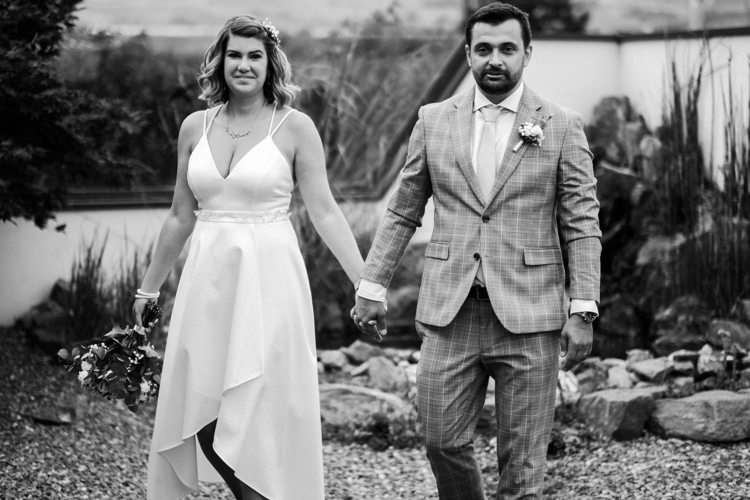Wedding couple in elegant attire holding hands while walking through garden setting.