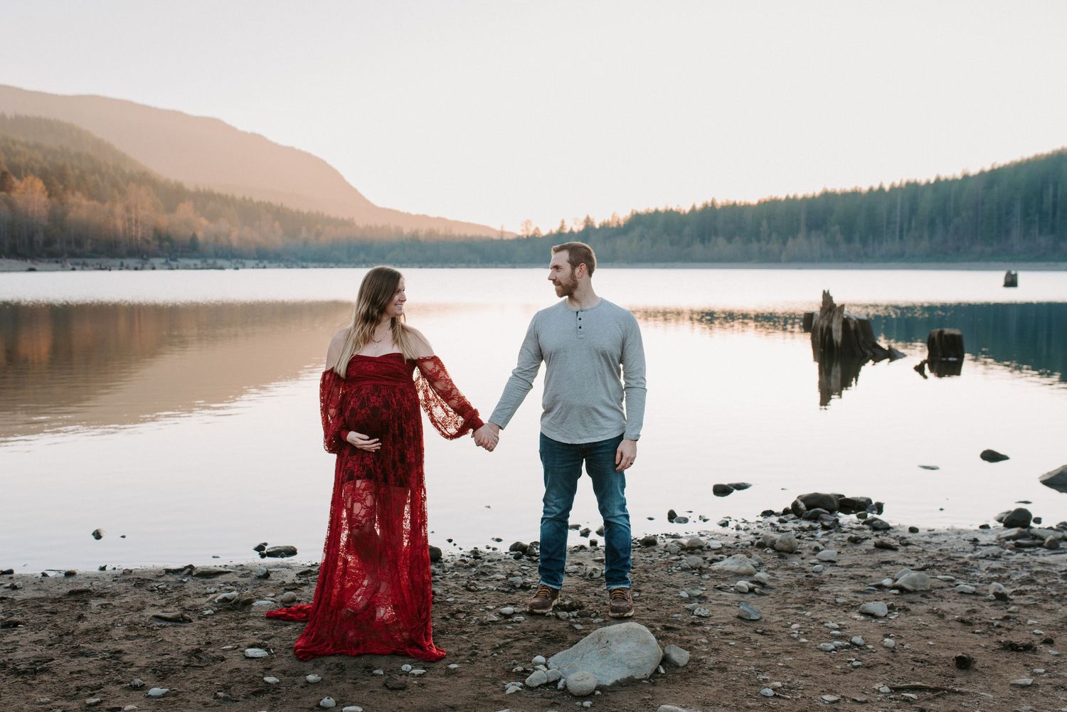 Couple walking along rocky shoreline near mountain lake at sunset.
