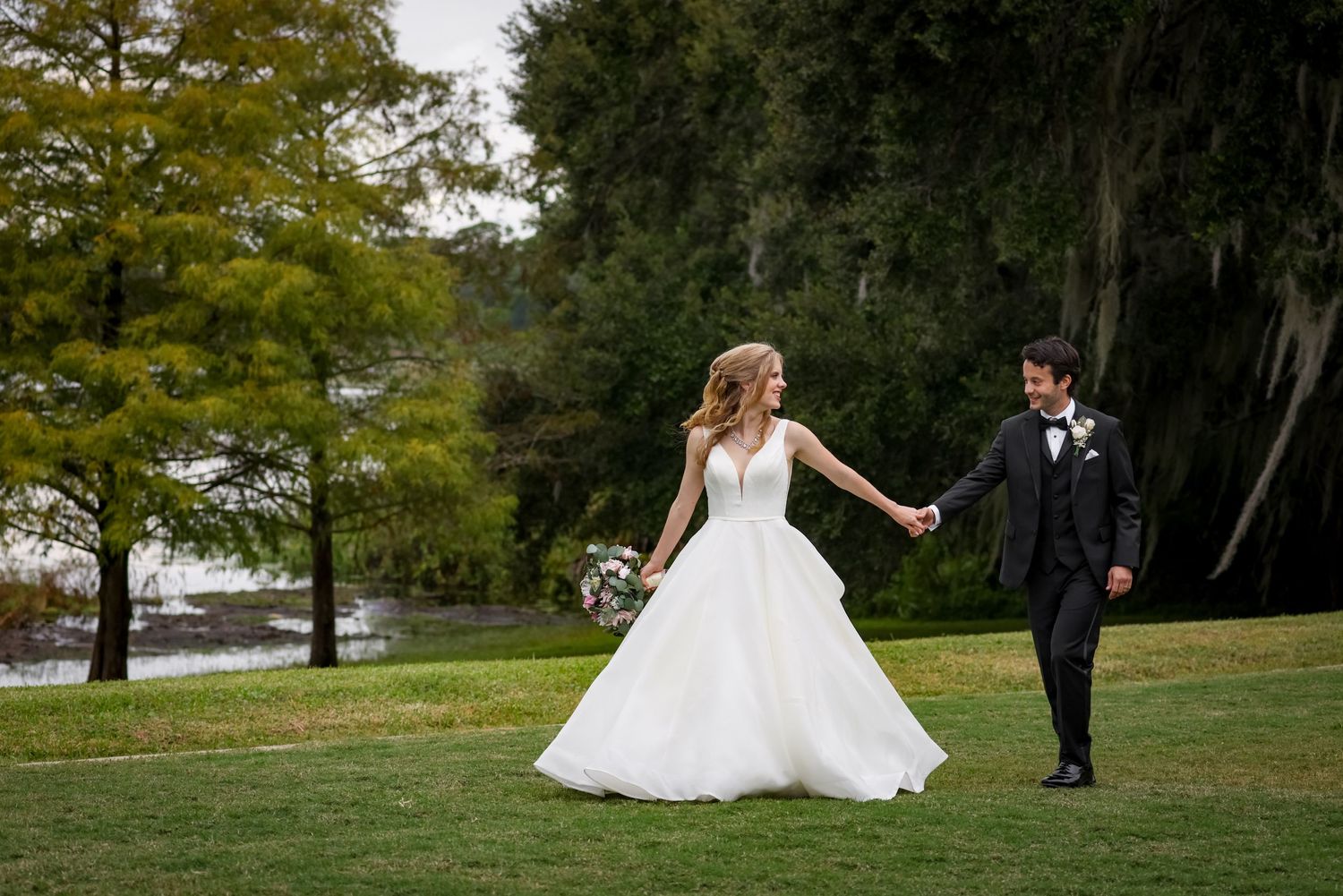Romantic wedding photo of couple walking on manicured lawn by waterfront.