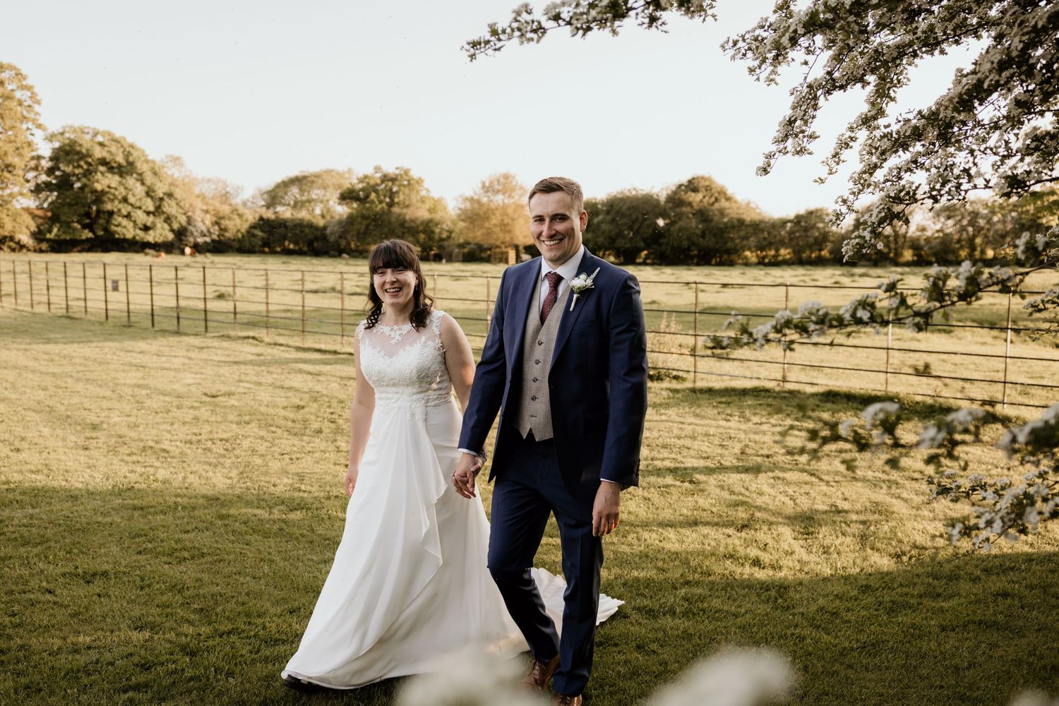 A wedding couple walks together in a golden sunlit field during their outdoor countryside celebration.