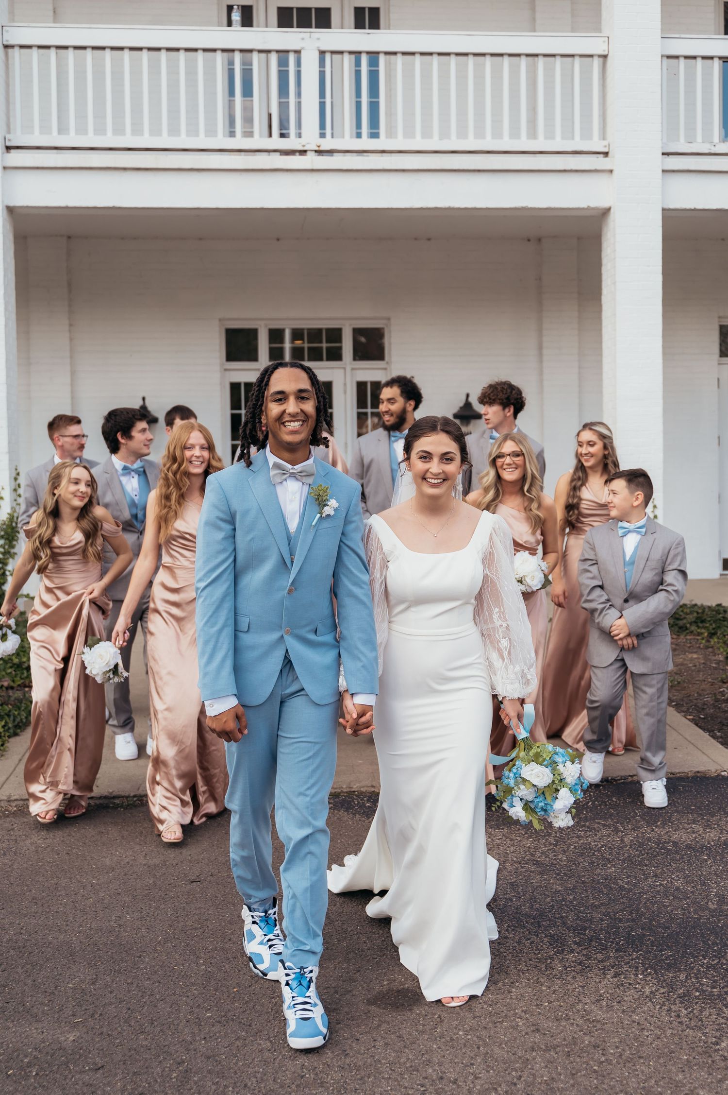Wedding party in pastel attire poses on white porch with couple in blue suit and white dress.