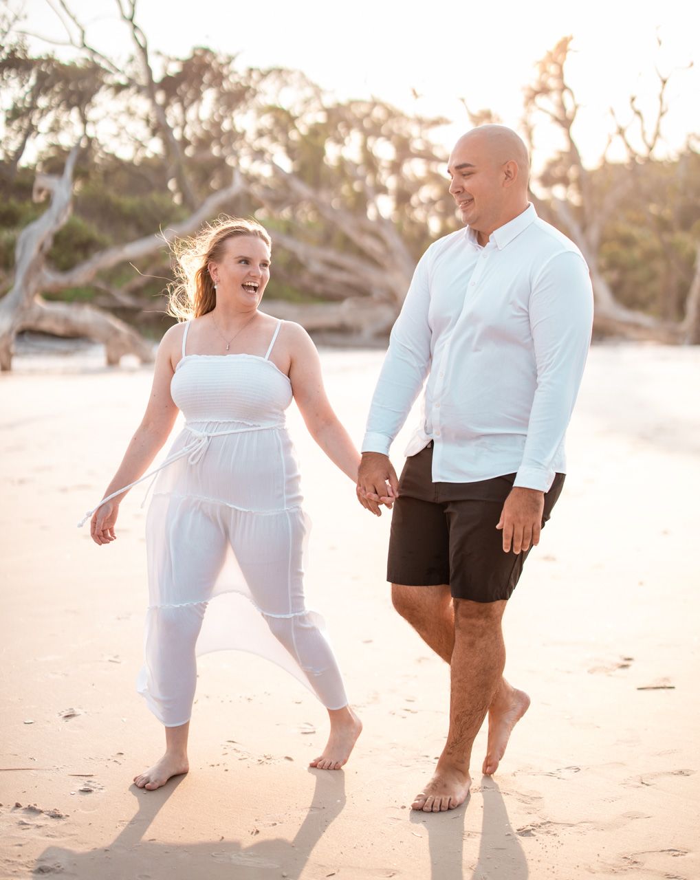Couple laughing together during sunset beach photoshoot wearing all white outfits.