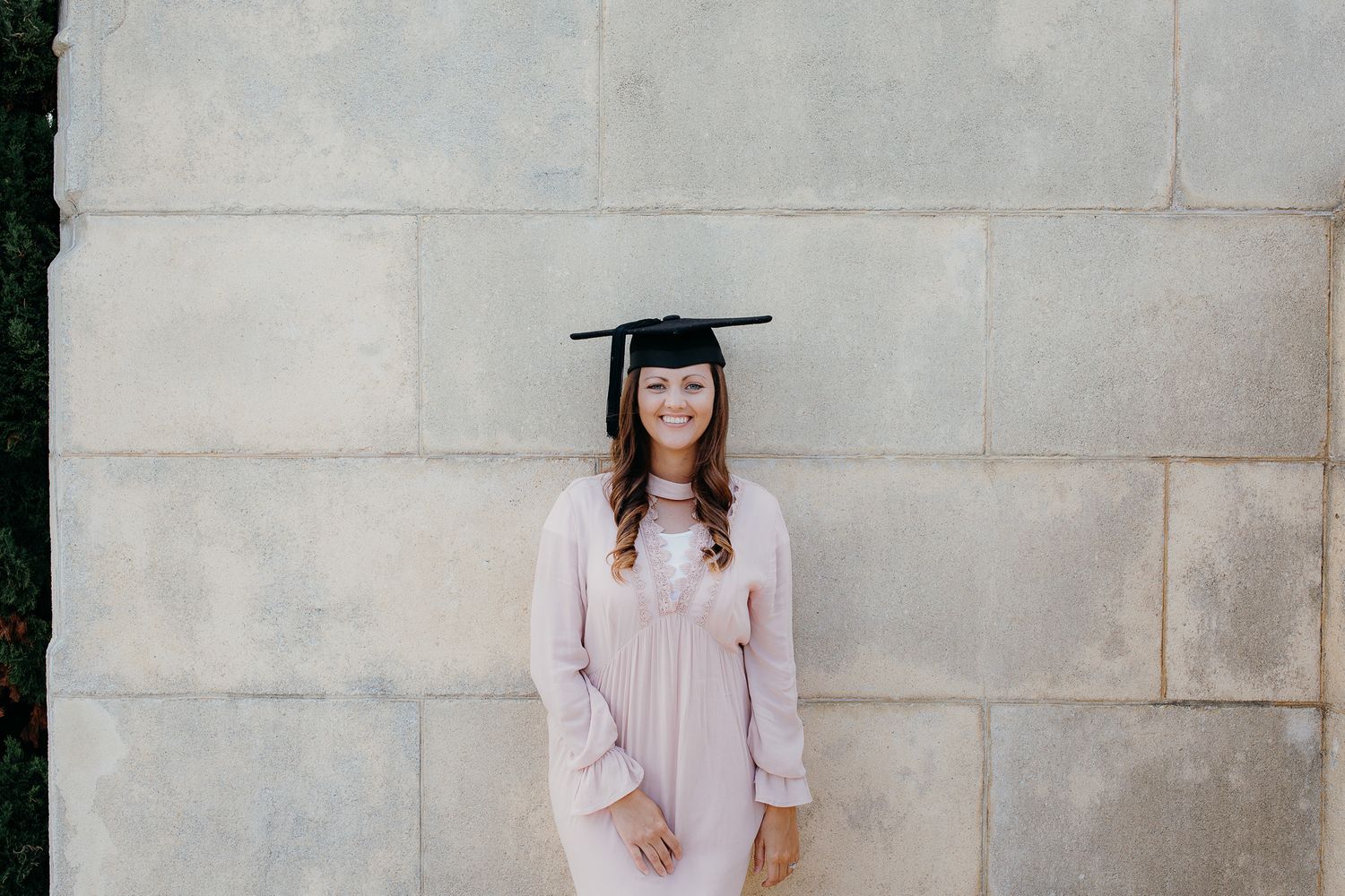 woman in pink dress wearing a graduation cap smiles at the camera while standing in front of a stone wall