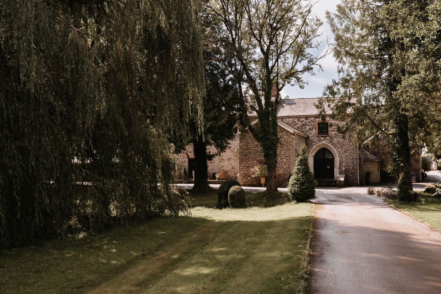 Stone church or building entrance with willow trees and curved driveway on a sunny day.