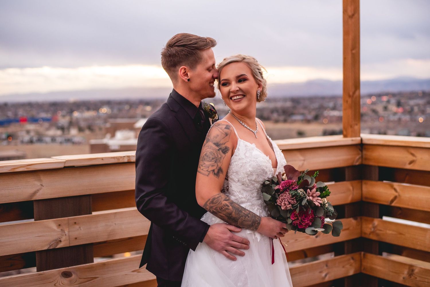 A wedding couple shares an intimate moment on a wooden deck overlooking a city landscape.