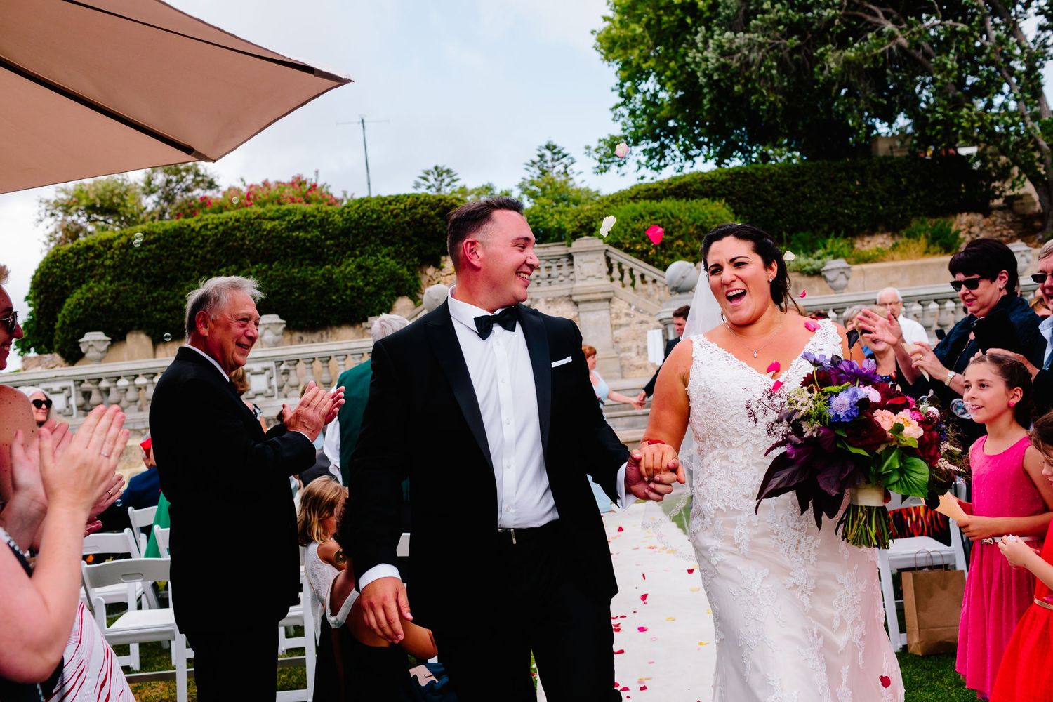 Wedding couple walks down outdoor aisle while guests applaud and celebrate.