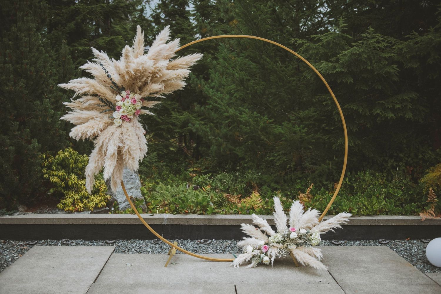 Modern wedding arch with dried pampas grass decorations against lush garden backdrop.