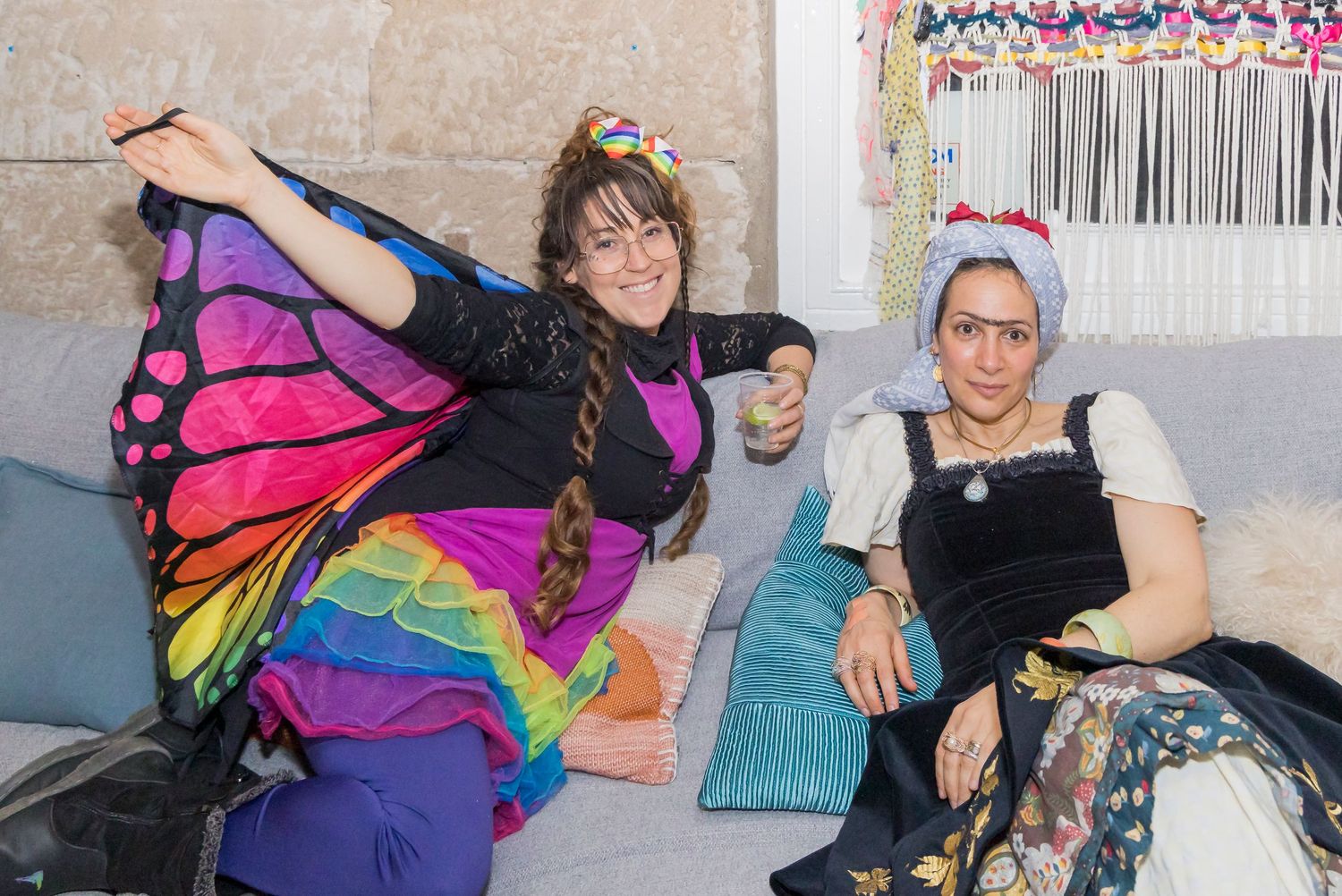 Person wearing colorful butterfly wings poses next to someone sitting on cushions.