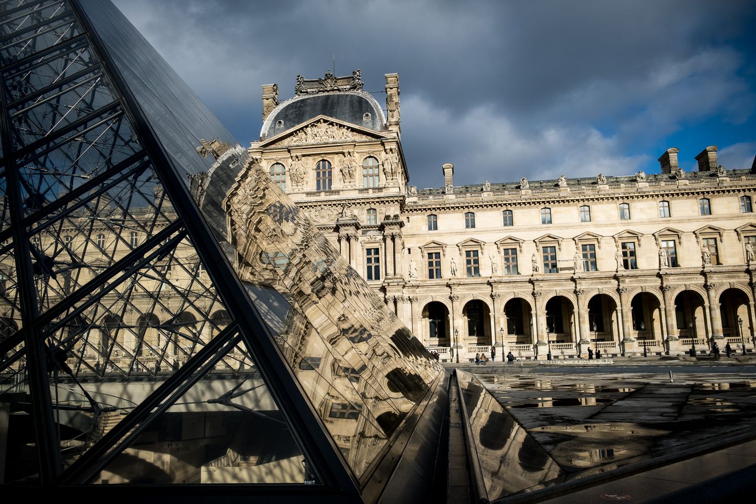 The Louvre Museum courtyard in Paris featuring the famous glass pyramid under dramatic cloudy skies.